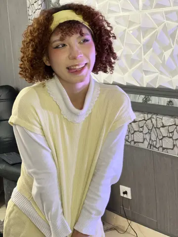 Jessiee_Belle with curly reddish hair and a yellow headband, wearing a pale yellow ribbed sweater and smiling.