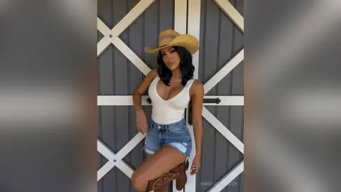 Stylish woman in a white ribbed tank top, denim shorts, and cowboy hat posing by a rustic grey barn door.