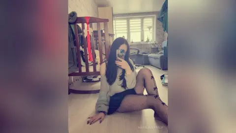 MarieJosePhilip sits on the floor wearing a grey sweater and black fishnet stockings, posing for a mirror selfie in her room.