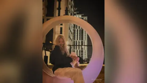 Blonde woman in a black blazer and beige trousers relaxing in a circular swing chair at a night rooftop lounge.