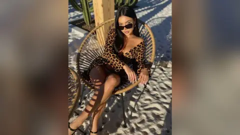 Stylish woman in a leopard print dress and sunglasses sitting in a wicker chair on a sunny beach.