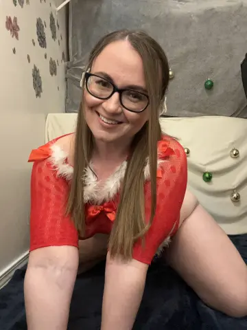 Smiling woman in a red and white festive holiday outfit sitting on a bed in a cozy indoor setting.