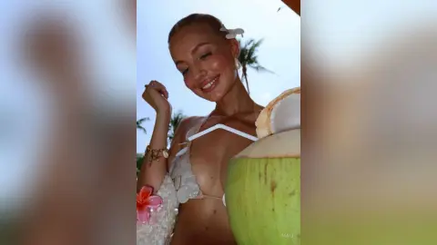 Smiling blonde woman in a white floral bikini holding a fresh coconut on a sunny tropical beach.