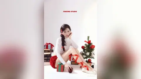 Young woman in white turtleneck and red skirt sitting by a Christmas tree with festive gift boxes.