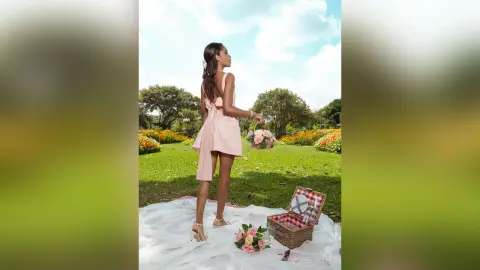 Young woman in a pink summer dress holding a flower basket in a sunny meadow with a picnic blanket.
