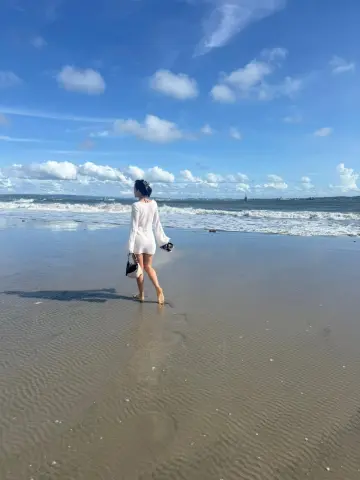 Woman in a white long-sleeve dress walking on a sunny beach under a blue sky.
