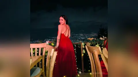 Smiling woman in elegant red evening gown posing on a balcony overlooking city lights at night.