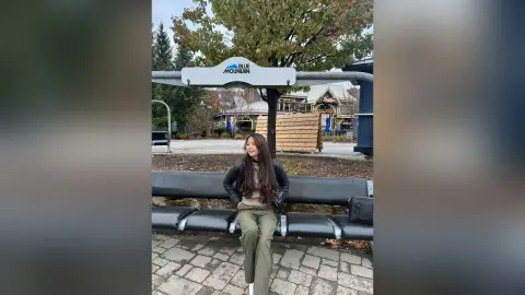 Smiling woman in a black jacket and olive pants sitting on a bench at Blue Mountain resort on an autumn day.