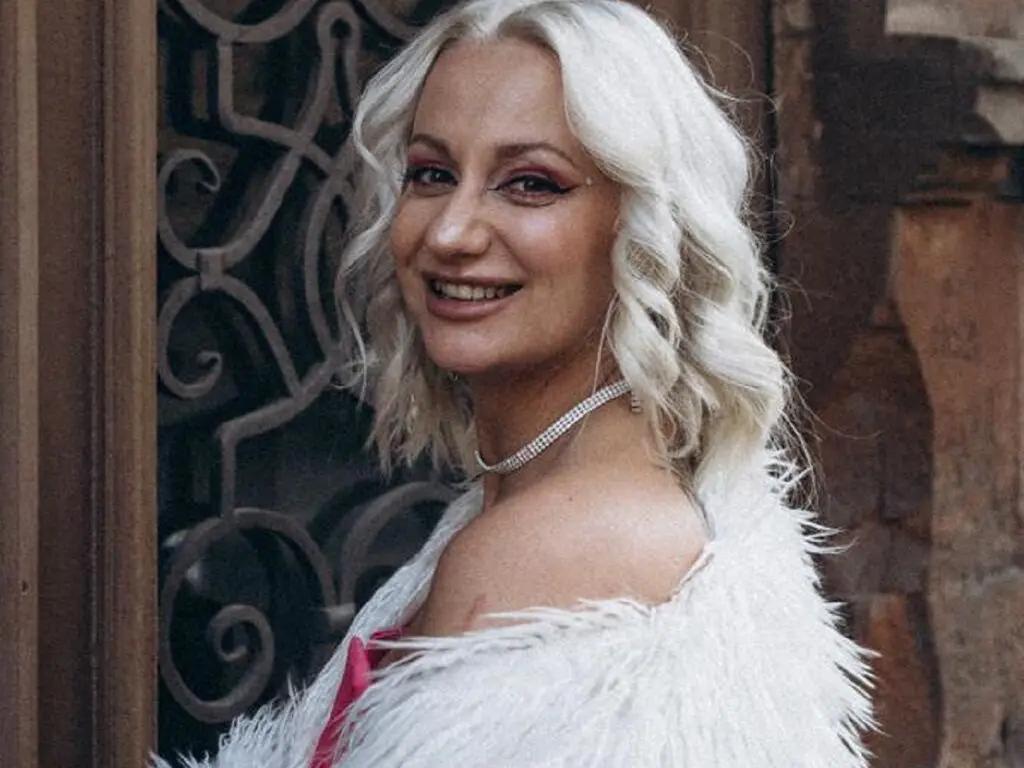 Smiling woman with wavy white hair wearing a white feathered outfit in front of an ornate metal gate.