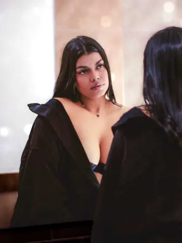 Young woman with long dark hair wearing a black robe looking into a mirror in a warm, moody indoor setting.