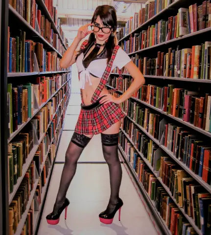 MistressMadelyn poses in a library wearing a red plaid skirt, white crop top, black tie, and black thigh-high stockings with pink platform heels.
