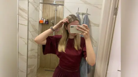 Young woman in a burgundy blouse taking a mirror selfie in a modern marble bathroom.