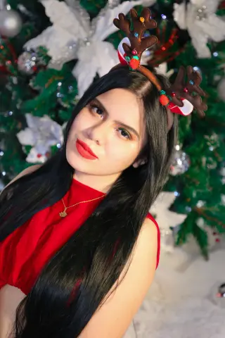 Young woman in a red dress and reindeer headband smiling in front of a festive Christmas tree.