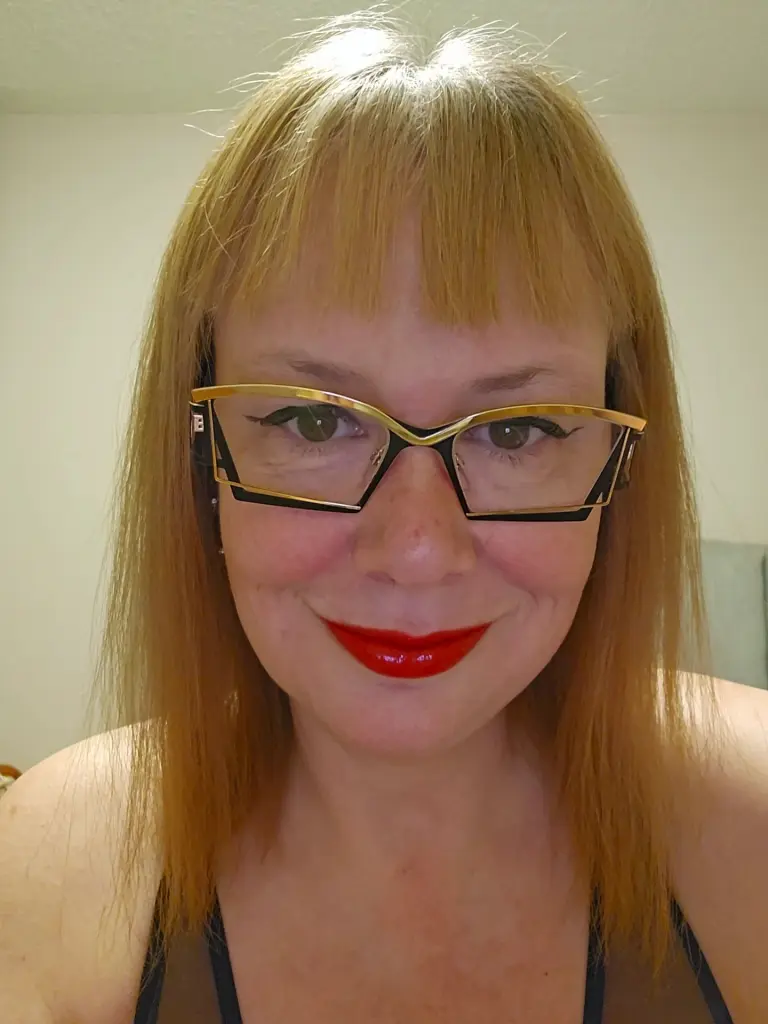 Smiling woman with blonde bangs and glasses wearing a pearl necklace and light blue strap top indoors.
