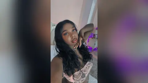 Young woman with long wavy hair wearing a pink lace bralette in a soft, dreamy bedroom setting.