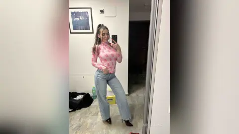 Young woman in a pink patterned turtleneck and light blue jeans taking a mirror selfie in a modern room.
