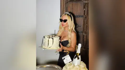 Blonde woman in a black dress celebrating with white cake and champagne in a luxury indoor setting.