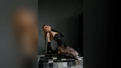 Elegant woman in black lace bodysuit and long gloves posing on a checkered platform against a dark background.
