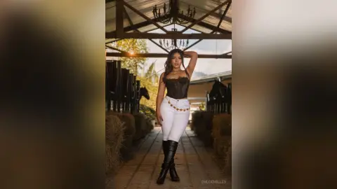 Stylish woman in a black corset and white jeans posing confidently in a rustic wooden stable setting.