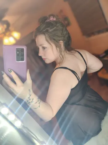 Young woman in a black slip dress taking a moody mirror selfie in a dimly lit room.