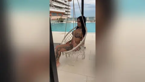Tattooed woman in a leopard print bikini relaxing on a hanging chair by a luxury rooftop pool.