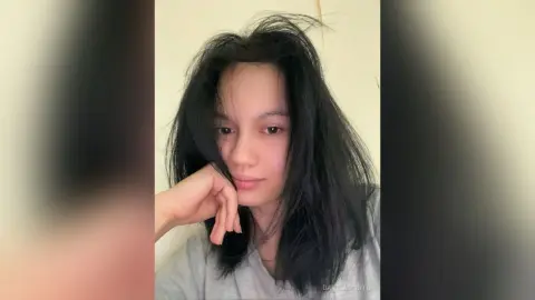 Young woman with long dark hair wearing a grey t-shirt in a minimalist indoor setting with a calm mood.