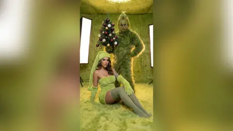 Woman in green holiday outfit sitting next to Grinch character by a decorated Christmas tree in a festive setting.