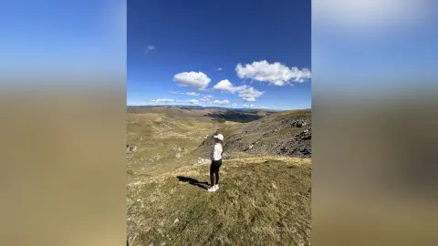 Adventurous woman in white top and black leggings standing on a grassy mountain ridge under a clear blue sky.