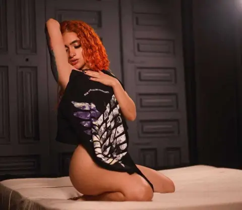Young woman with curly red hair wearing a black graphic t-shirt sitting on a bed in a moody indoor setting.
