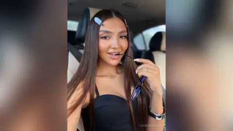 LeonidaAbdon poses in a car wearing a black top, holding sunglasses near her mouth with long brown hair and hair clips.