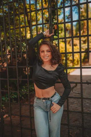 Smiling young woman in a black long-sleeve crop top and blue jeans leaning against a black metal fence outdoors.