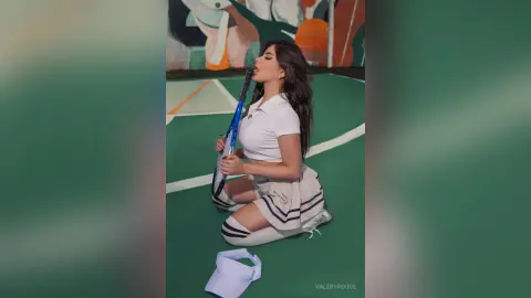 Young woman in white tennis outfit sitting on a green court holding a blue racket with a playful mood.