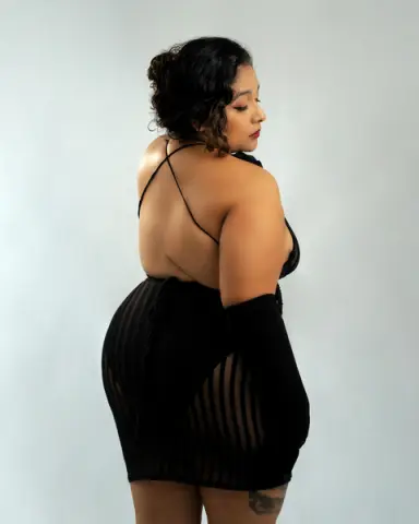 Curvy woman in a black backless mini dress posing against a plain studio background with a confident mood.