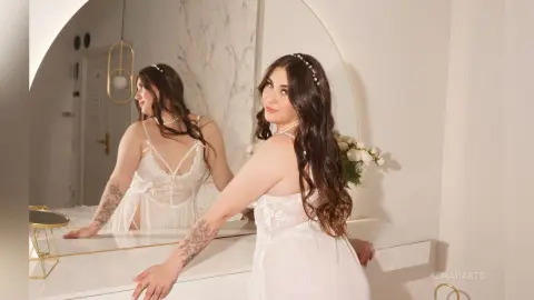 KeiraHarts poses in a white lace slip, looking back over her shoulder while showing off her long wavy hair and arm tattoos in front of a mirror.