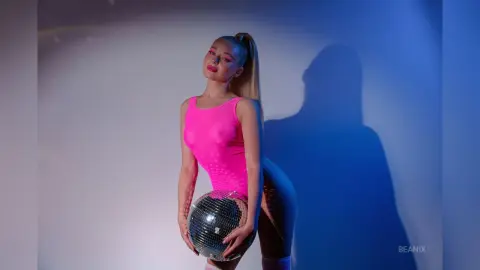 BeaNix poses in a bright pink bodysuit while holding a disco ball against a blue-lit background.