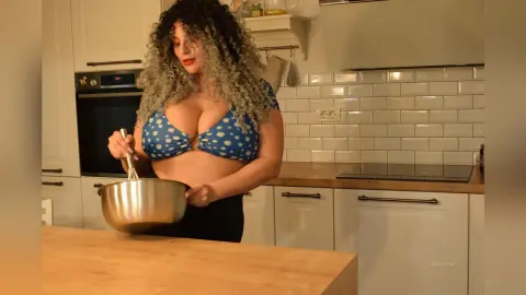 Curvy woman with curly hair in a blue floral bikini top cooking in a bright modern kitchen.