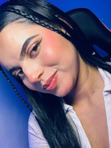 Smiling woman with long black braided hair and winged eyeliner wearing a white shirt against a blue background.