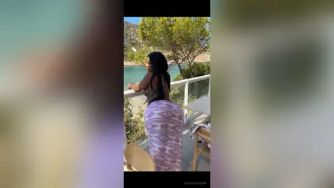 Woman in a patterned white and purple maxi dress relaxing on a balcony overlooking a scenic lake and mountains.