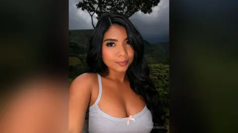 Beautiful woman with long wavy hair in a grey tank top posing against a lush green mountain landscape.