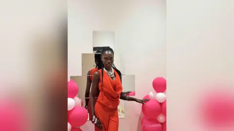 Smiling Black woman in a vibrant red jumpsuit posing in a bright room with pink balloons.