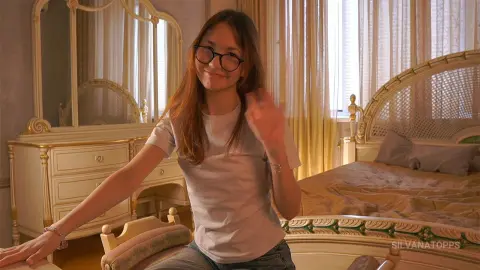 Smiling young woman with glasses in a white t-shirt sitting in a bright, vintage-style bedroom.