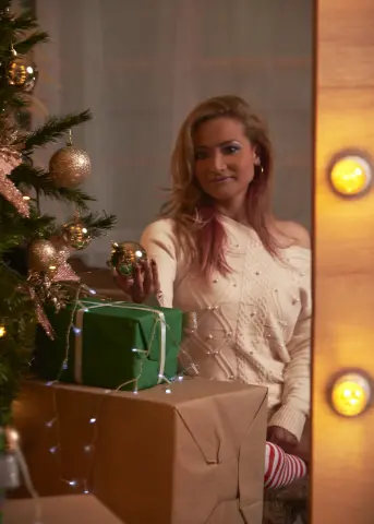 Smiling woman in a white knit sweater sitting by a decorated Christmas tree with wrapped gifts in a cozy mood.