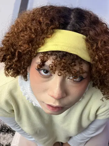 Jessiee_Belle looking up at the camera with curly reddish-brown hair and a yellow headband, wearing a pale yellow ribbed top.