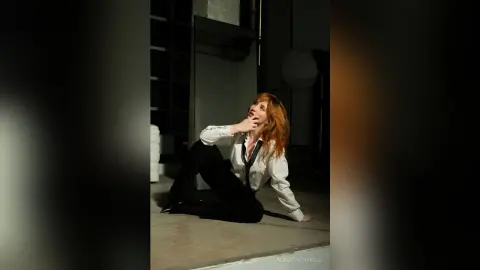 AlbertaCorrell sits on the floor in a white shirt and black pants, looking up with her hand to her chin.