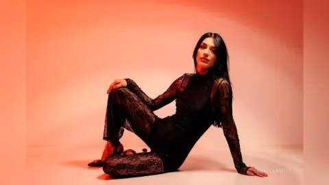 NerissaMist poses in a black lace bodysuit against a red background, looking at the camera with long dark hair.