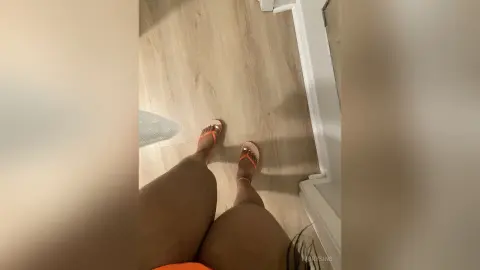 Close-up of a woman's legs wearing orange strappy sandals on a light wood floor in a bright room.