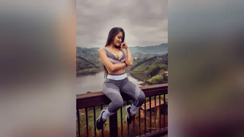 Young woman in grey yoga set sitting on a balcony overlooking a scenic mountain lake landscape.