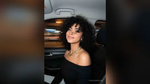Smiling young woman with curly hair wearing a black off-the-shoulder top sitting in a car at night.