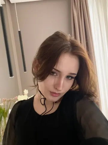 Young woman with brown hair wearing a black blouse and choker in a bright, modern room.
