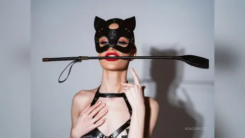 Model in a black leather cat mask and harness holding a whip against a grey studio background.